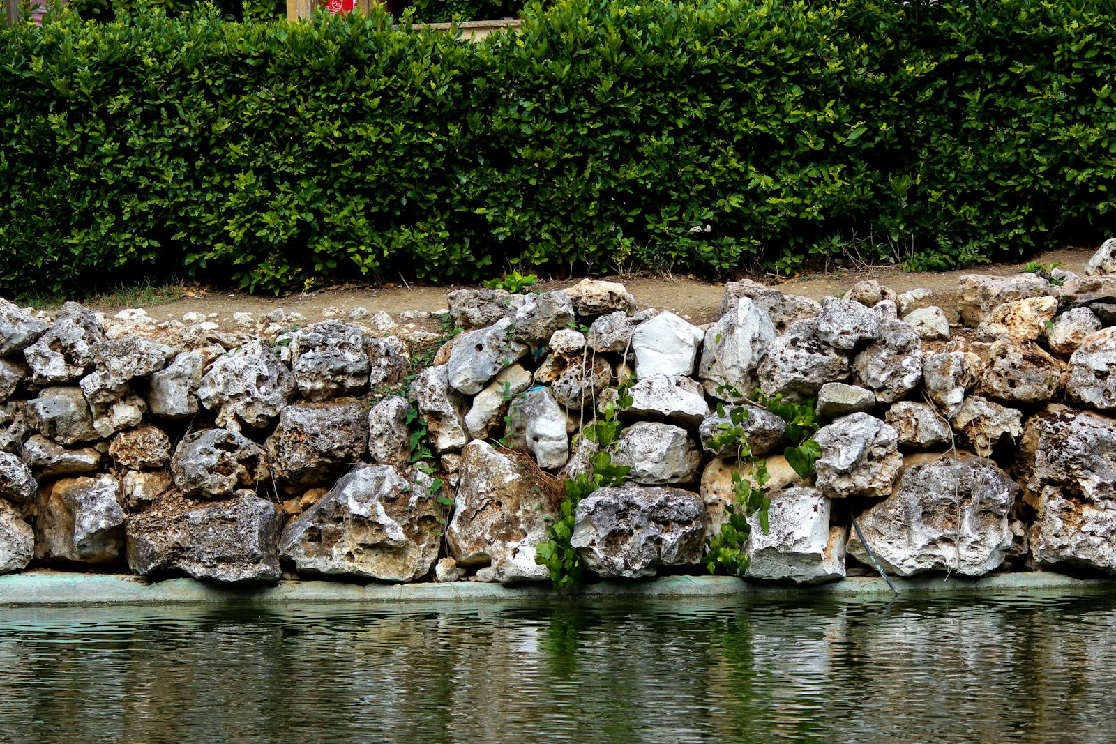 Tan stone retaining wall with a small water feature and trimmed hedges behind.